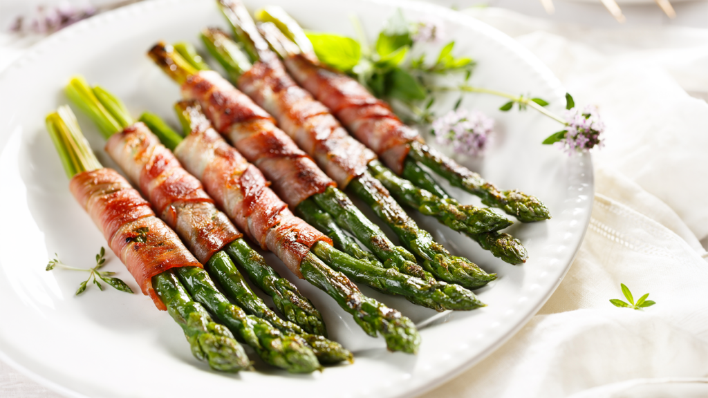 Tender grilled asparagus wrapped in prosciutto and finished with beurre blanc sauce, plated on a white dish with fresh herbs.