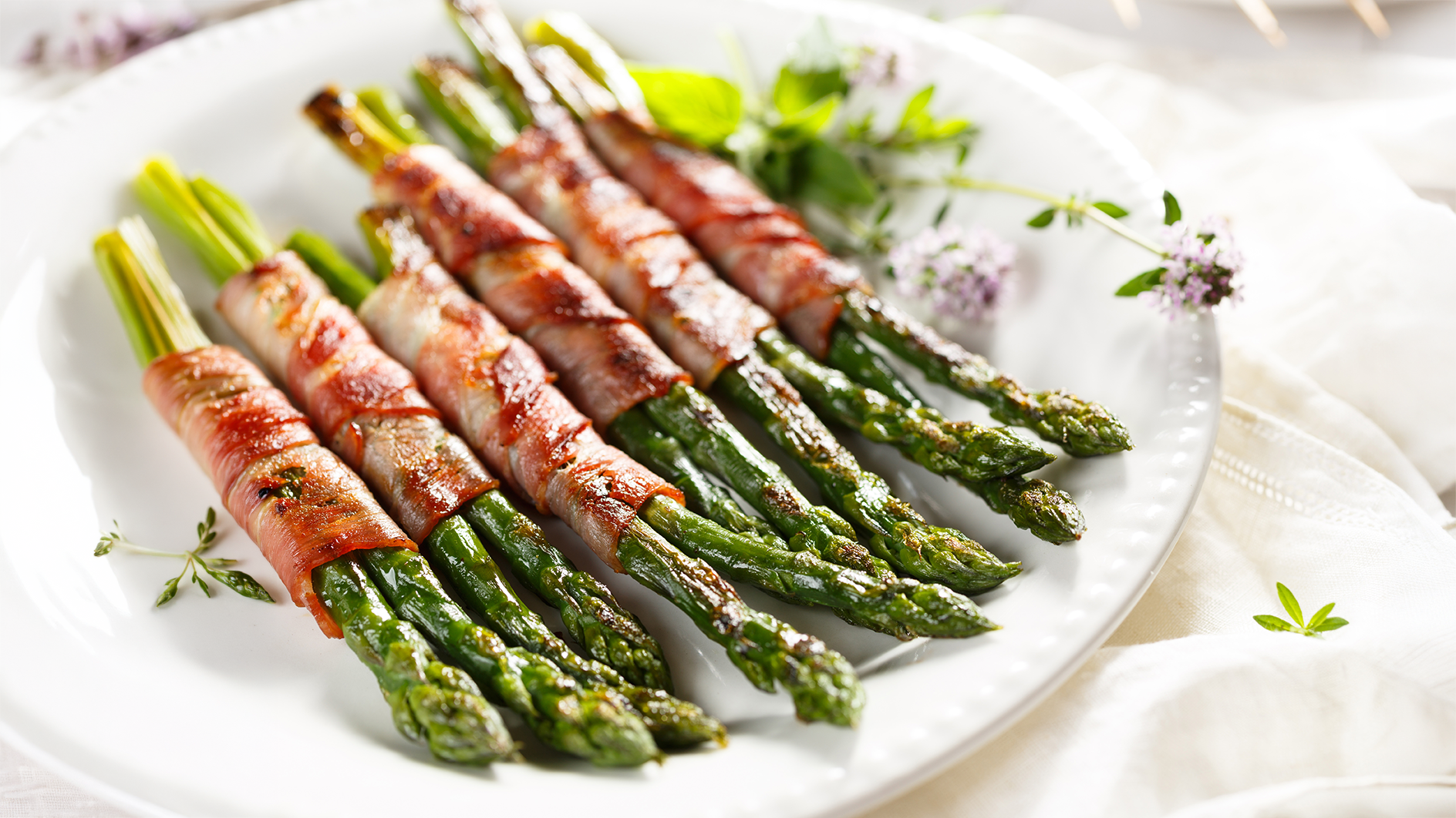 Tender grilled asparagus wrapped in prosciutto and finished with beurre blanc sauce, plated on a white dish with fresh herbs.