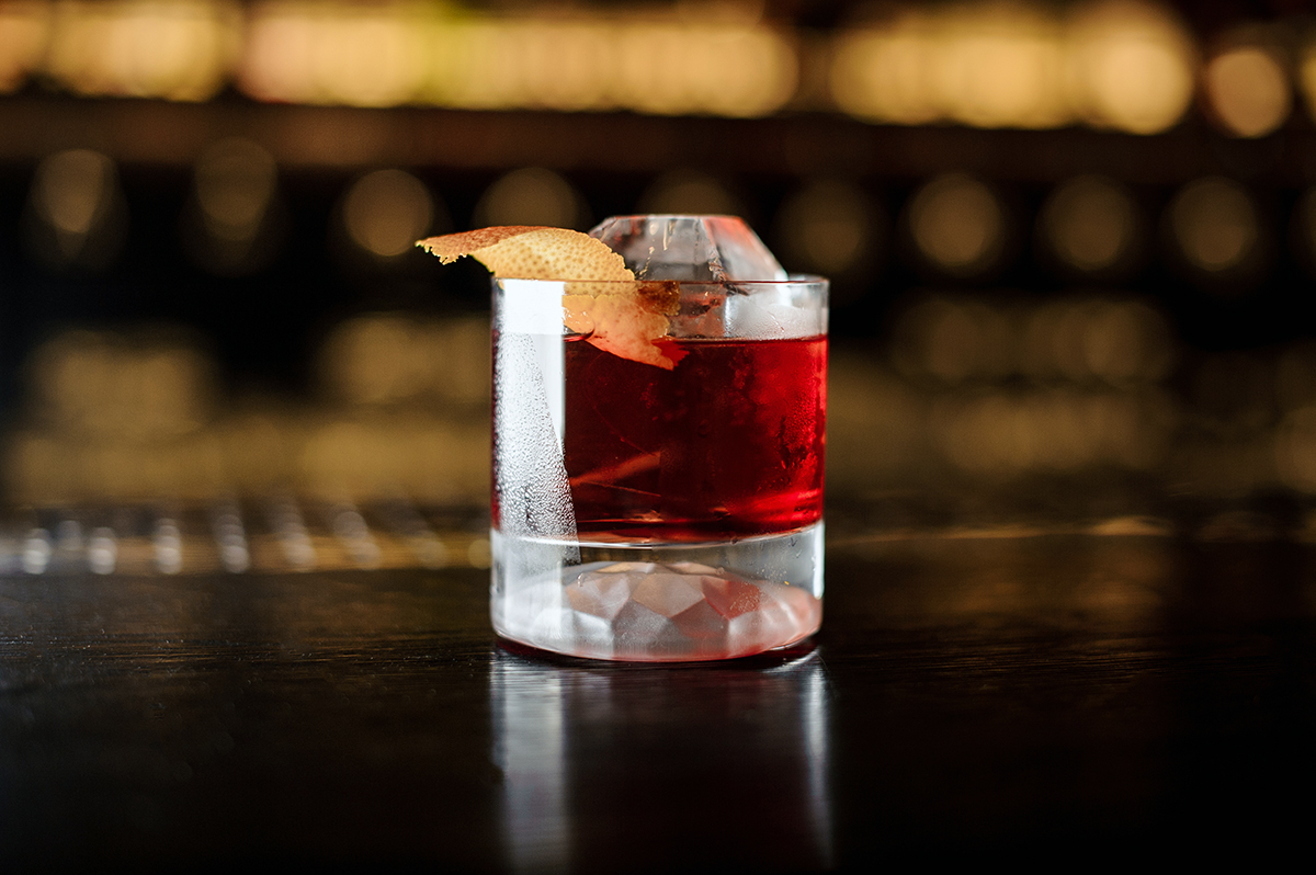boulevardier cocktail in rocks glass with big piece of ice and orange twist on wooden bar