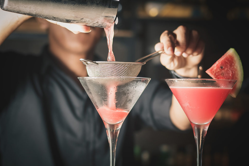 bartender double straining a watermelon gimlet into a martini glass garnished with watermelon slice
