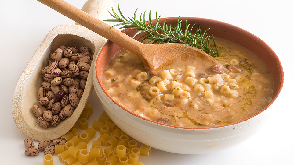 bowl of pasta fagipoli with wooden spoon in it and rosemary and scattered beans and pasta on the table